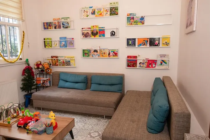 Duvar raflarında çocuk kitapları, kanepe ve oyuncaklı okuma köşesi